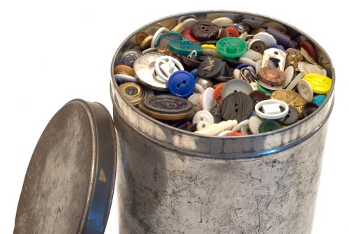 A metal storage drum filled with a variety of discarded plastic buttons of different sizes, shapes, and colours, including black, white, blue, green, yellow, beige, and brown, with some buttons featuring multiple holes and others with shank backs. The drum, which appears slightly worn with a scratched and tarnished surface, is positioned on a white background, with its removable lid placed nearby. The image is related to rubbish removal services, showcasing the type of waste that a company like Office Clearance Dalston might collect during a clearance in the Dalston area, highlighting the diverse range of small, mixed plastic waste items. The contents are densely packed, filling the drum close to the rim, and the lighting is even, emphasizing the different textures and colours of the buttons, which contrast against the metallic surface of the container, illustrating typical rubbish materials encountered in office or domestic clearance work within London’s East End.