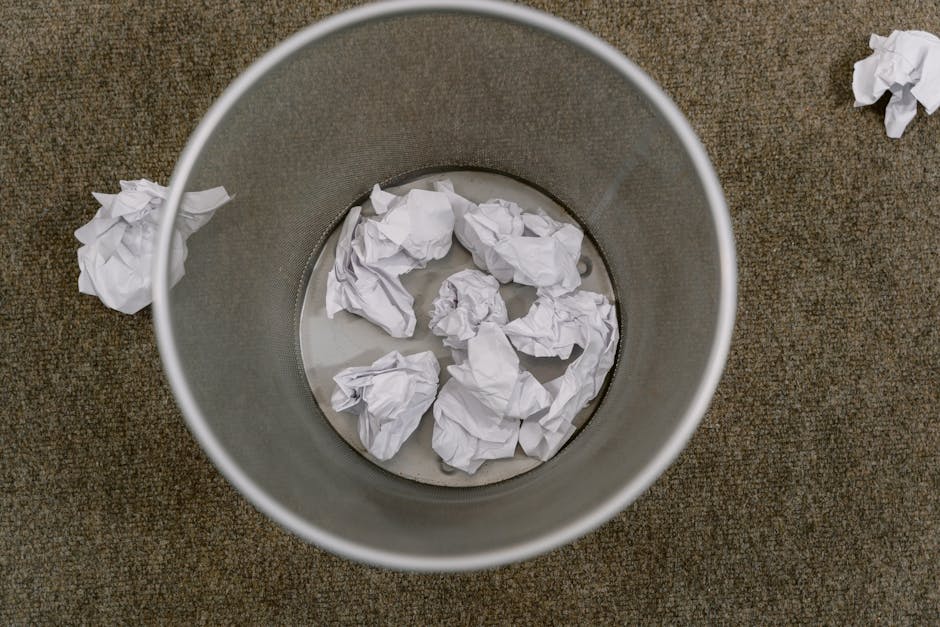 An overhead view of a cylindrical metal waste bin placed on a brown textured carpeted floor. The bin contains several crumpled white sheets of paper, some of which are partially submerged in a small a