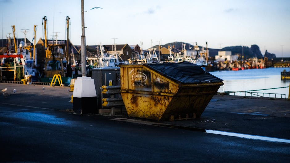 In the foreground, there are two large orange waste containers, both showing signs of dirt, grime, and weathering, with one container filled with residuals and the other appearing empty. Attached to t