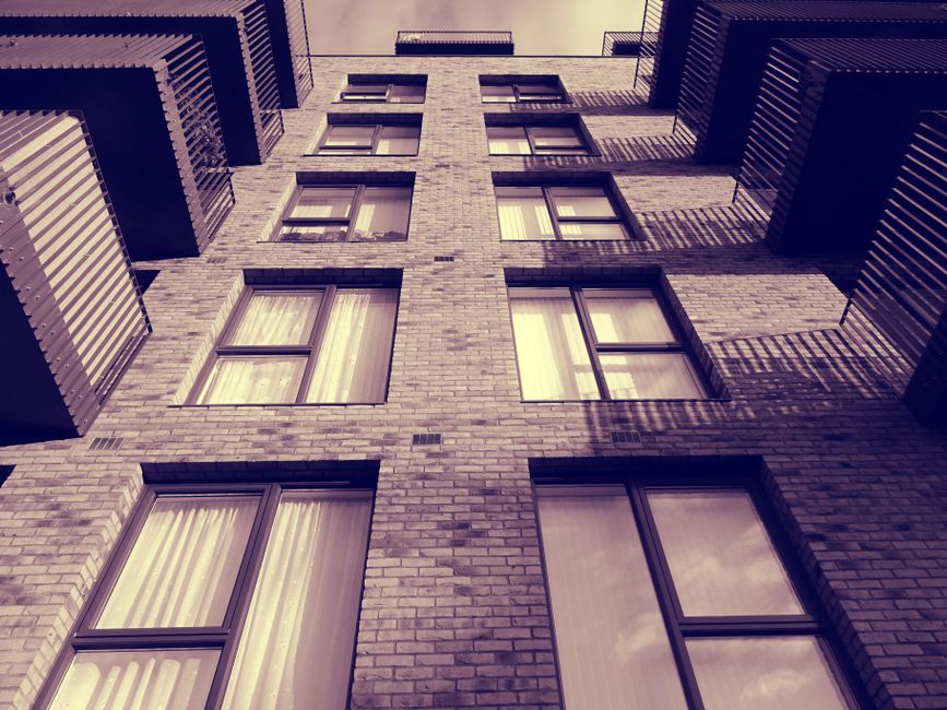 A tall multi-storey residential building constructed with light-colored bricks, featuring several rectangular windows with dark frames. Some windows have drawn curtains or blinds, and the building's v